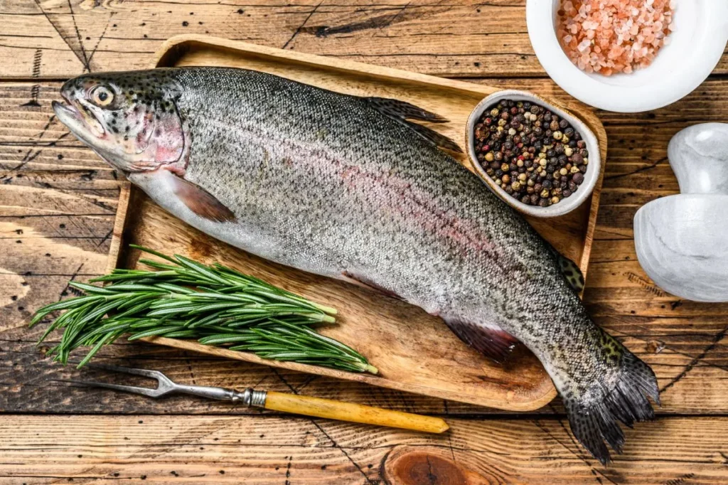 Steelhead Trout Recipe - Fresh whole trout placed on a wooden tray, surrounded by rosemary sprigs, a bowl of mixed peppercorns, and Himalayan pink salt on a rustic wooden table