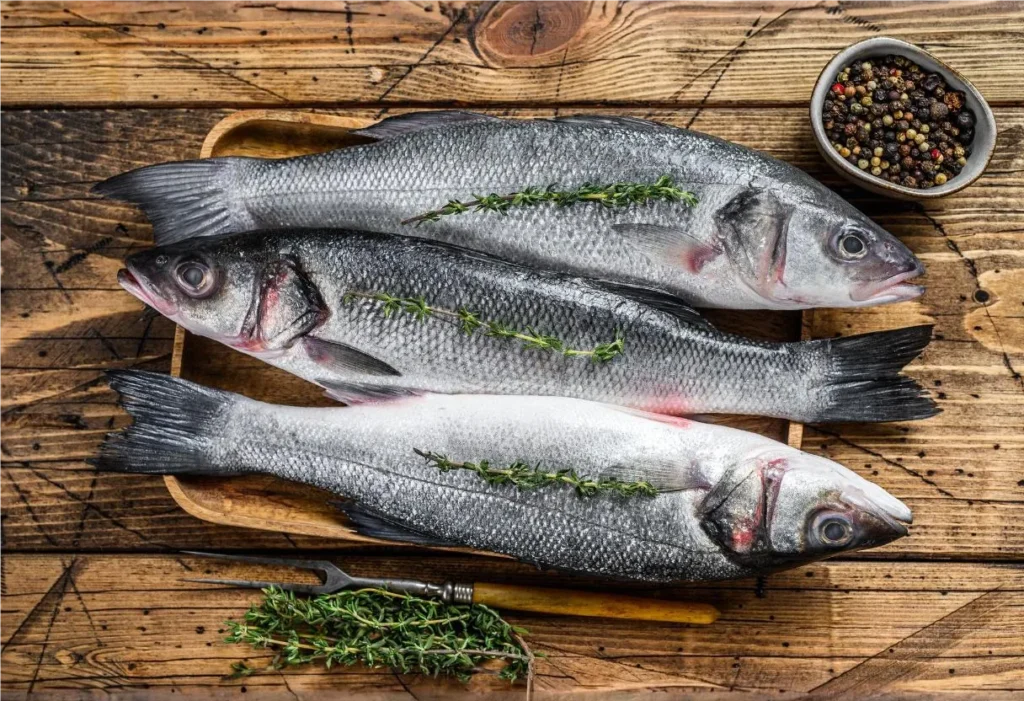 Three raw branzino fish garnished with herbs, ready to cook and eat, displayed on a wooden tray over a rustic table.