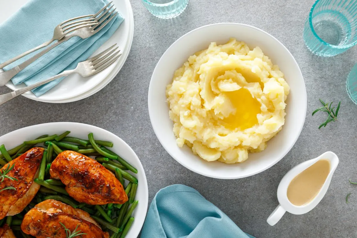 Mashed Potatoes and Gravy - A plate of roasted chicken breasts with green beans, served alongside a bowl of buttery mashed potatoes and a small gravy boat on a gray surface with blue napkins and glassware