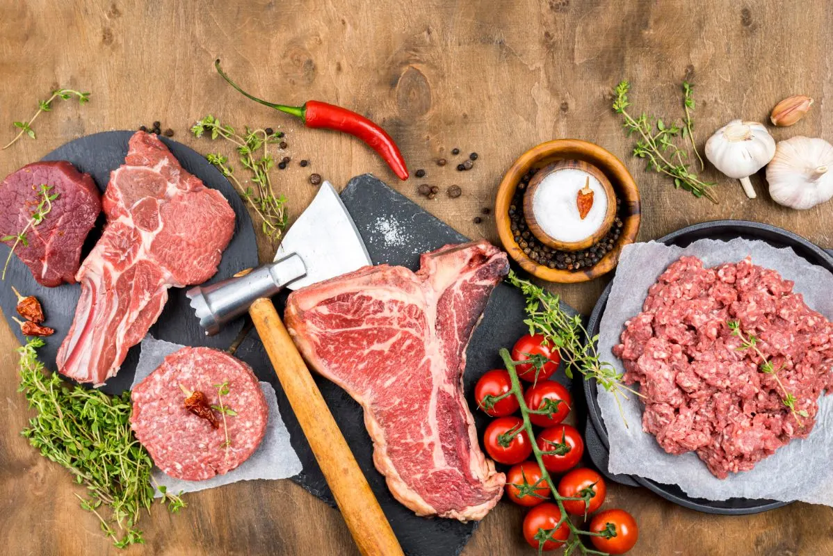 Best Meat for Chili - Various cuts of raw meat with herbs, garlic, tomatoes, and spices on a wooden surface