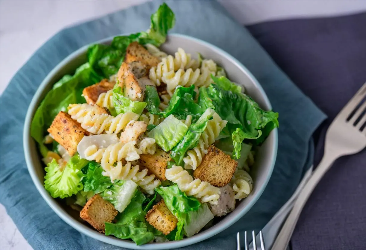 A bowl of Caesar pasta salad with rotini pasta, romaine lettuce, croutons, grilled chicken, and Parmesan cheese served in a white bowl.