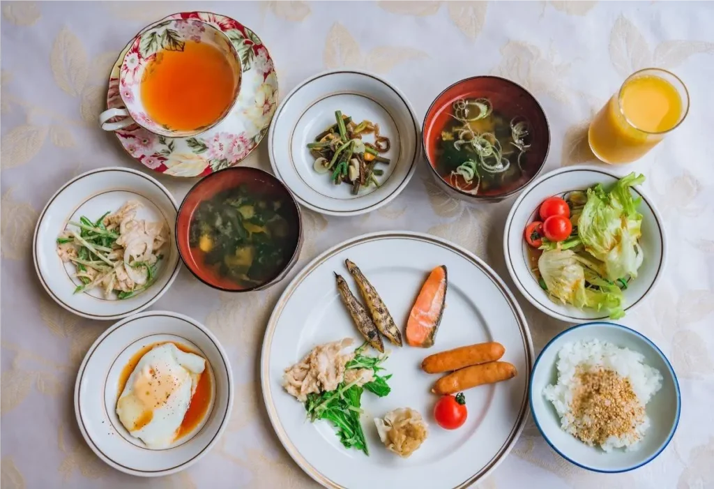 A traditional Japanese breakfast featuring grilled salmon, rice with sesame seeds, miso soup, sausages, side dishes, salad, tea, and orange juice served on a table.