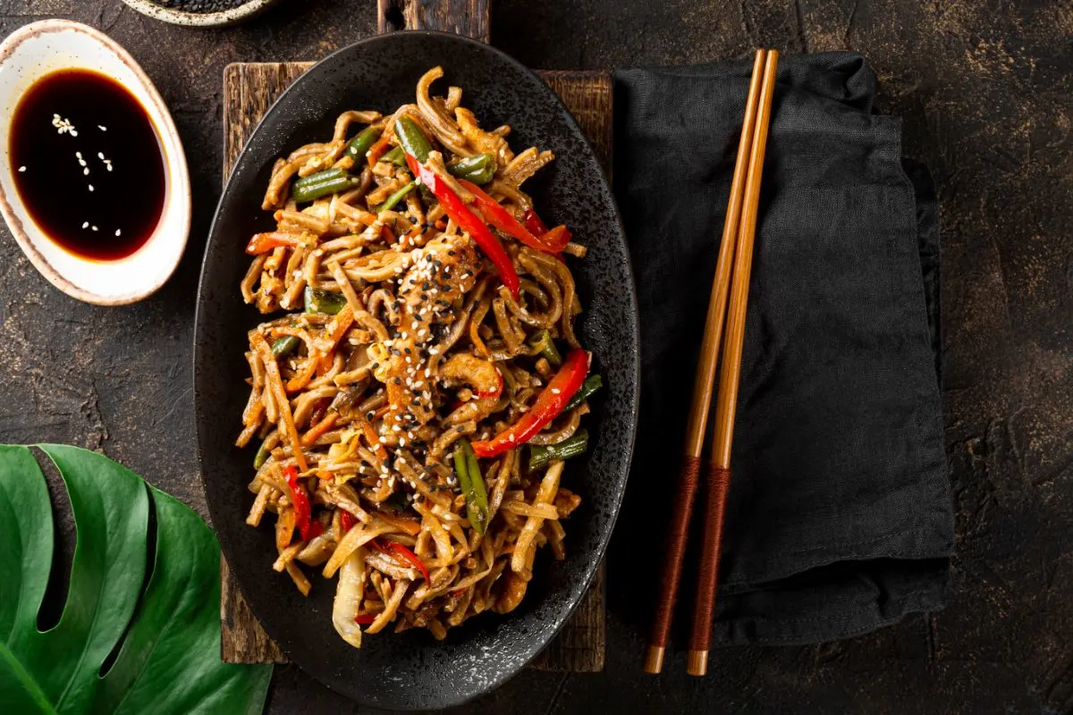 Hibachi Noodle Recipe - A black plate filled with stir-fried noodles, chicken, red bell peppers, green beans, and mushrooms, garnished with sesame seeds, served alongside chopsticks on a dark rustic background with a small bowl of soy sauce.