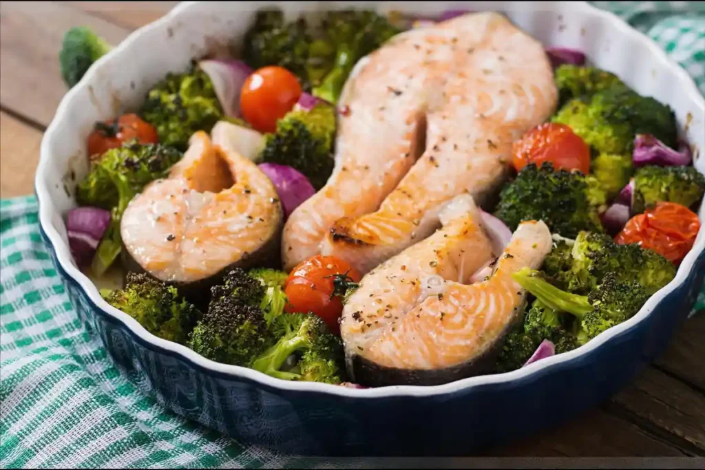 A baked salmon steak served with roasted vegetables, including broccoli, carrots, and cherry tomatoes, on a white plate.