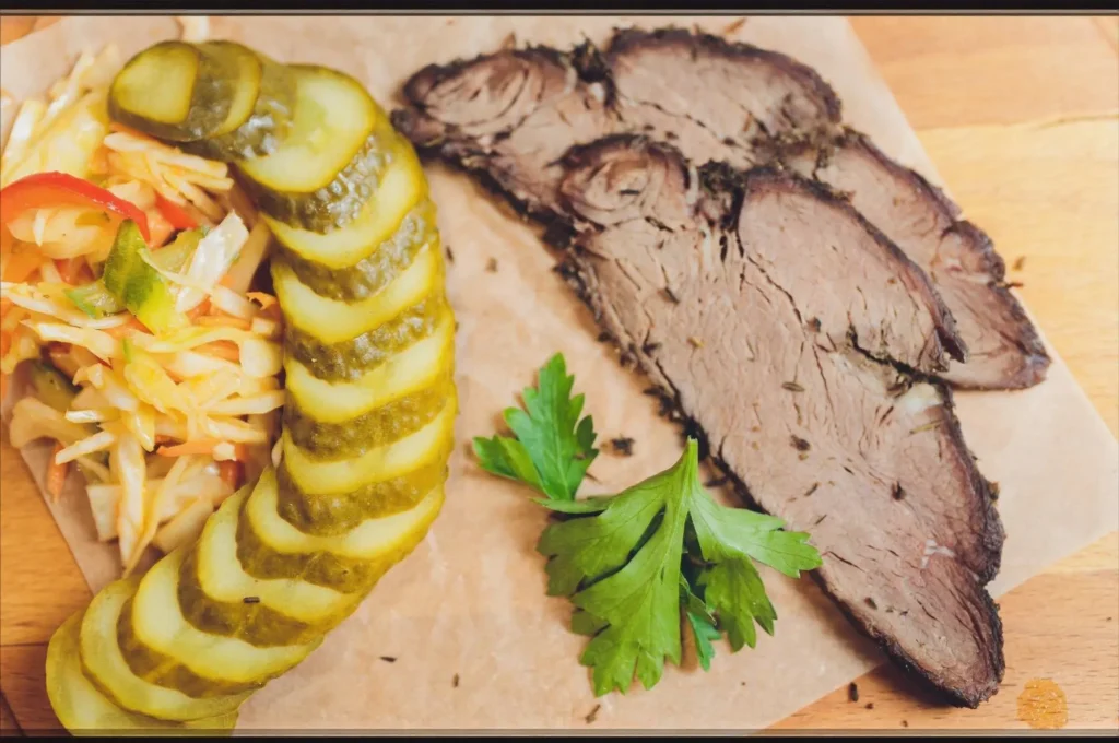 Brisket Sandwiches - A top-view photo of a roast brisket salad with sundried tomatoes, arugula, tomato slices, and cucumber, served on a white plate against a grey background.